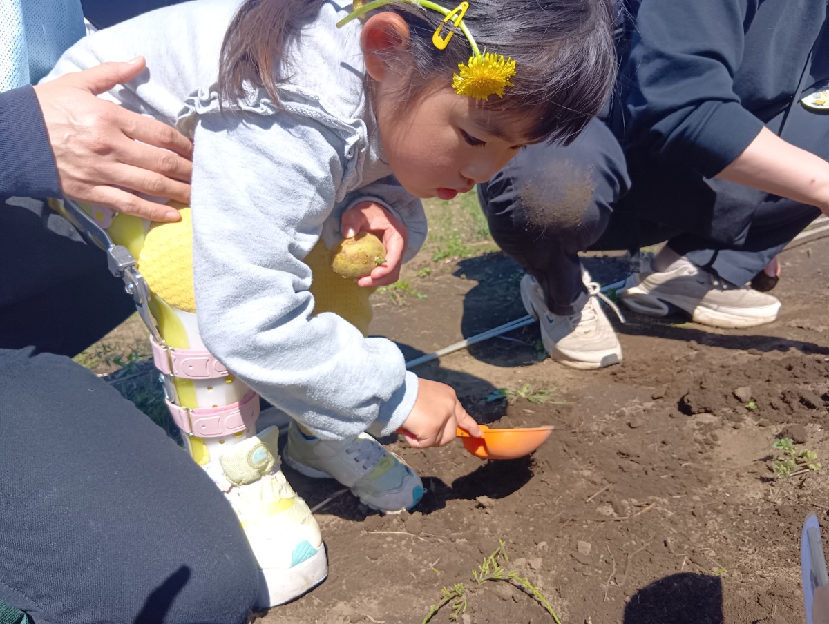 足に補助器具をつけた子どもが、後ろから大人に支えられて、片手に種芋、もう一方の手にはシャベルを持ち、畑の土をじっと見つめている。