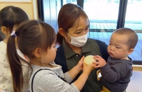 年上児が、保育者に抱っこしてもらっている年下児に玉ねぎを持っていき、年下児は玉ねぎに手を伸ばしている。