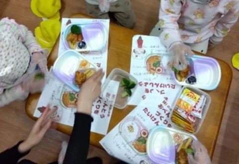 ビニール手袋を着けた子どもたちが、大人と一緒に、盛り付けの見本を見ながら弁当を盛り付けている。