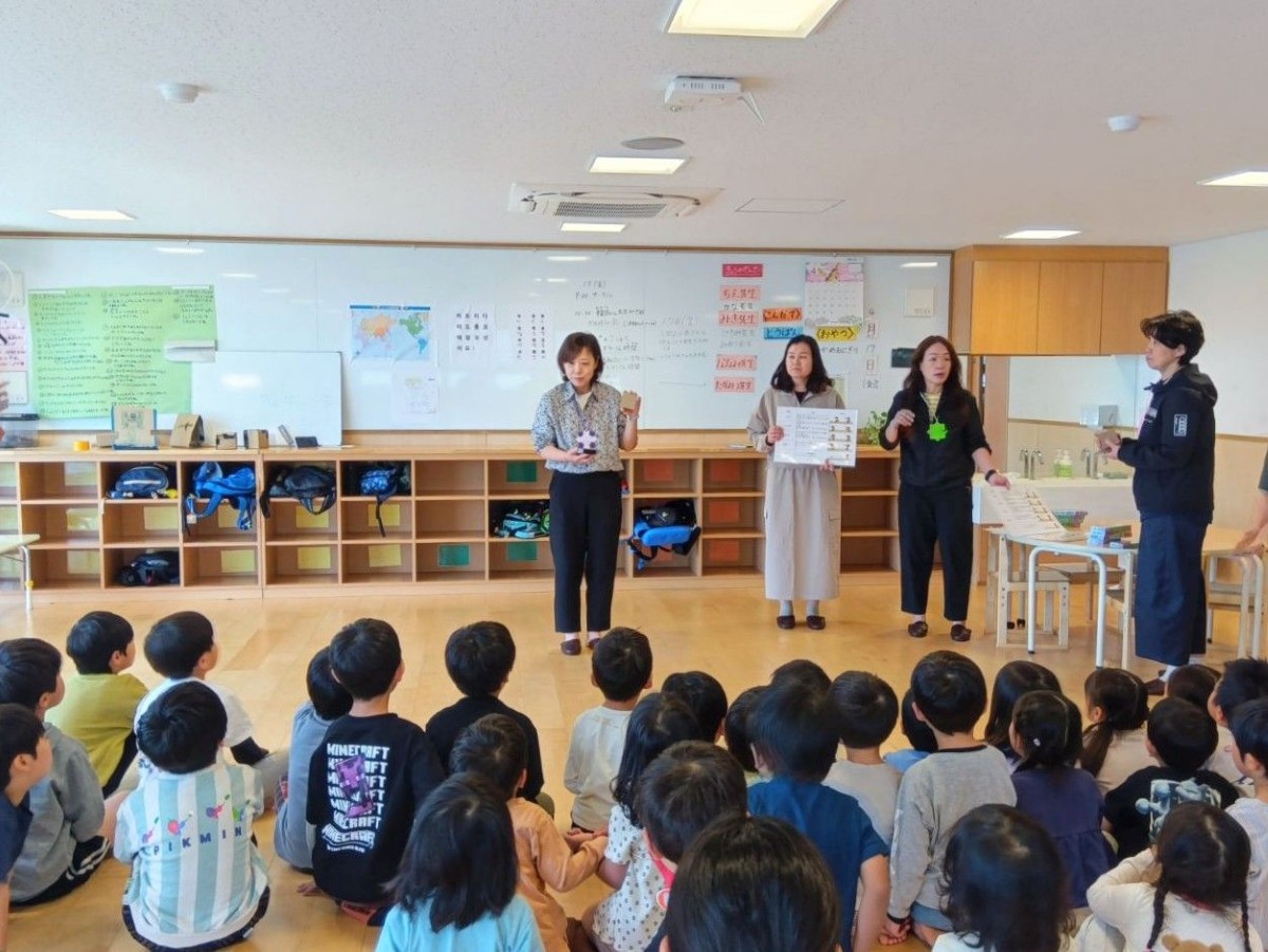 室内で、四人の大人が玩具とボードを持って子どもたちの前に立ち、子どもたちに話をしながら玩具をみせている。