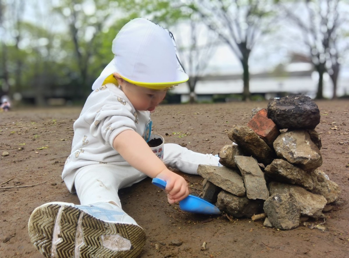 地面に座っている一人の子どもは、片手に土の入った器を持ち、もう一方の手にはシャベルを持って、積み上げられた石の周囲の土をすくおうとしている