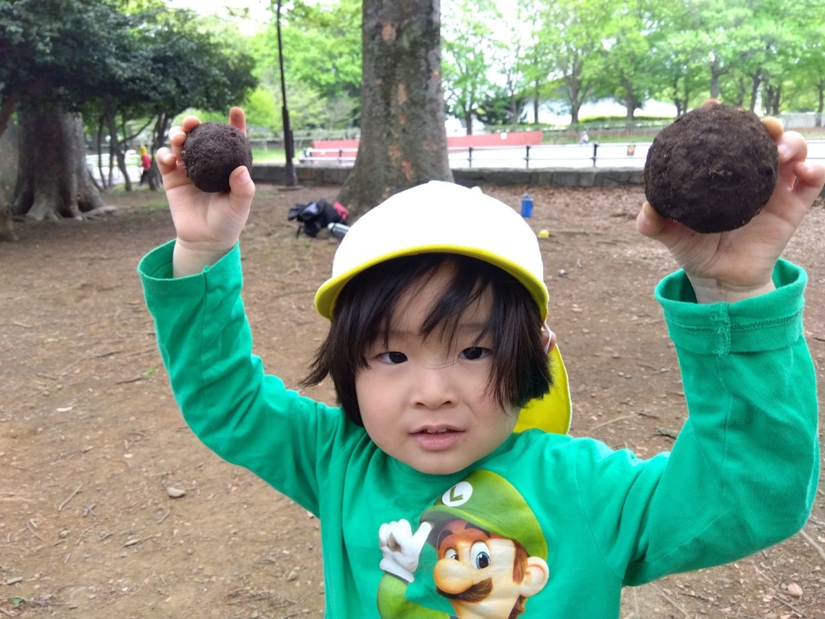 一人の子どもが、出来上がった2つ泥団子を左右の手に持ち、頭の高さまで上げて、カメラに向かって見せている。