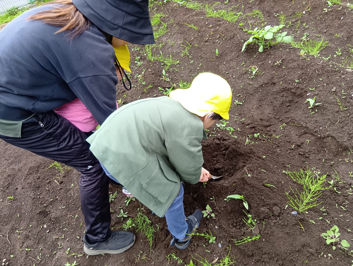 一人の子どもがスコップで土を掘っている。隣には子どもを抱えた大人がおり、隣の子どもと同じ姿勢で土づくりを手伝っている。