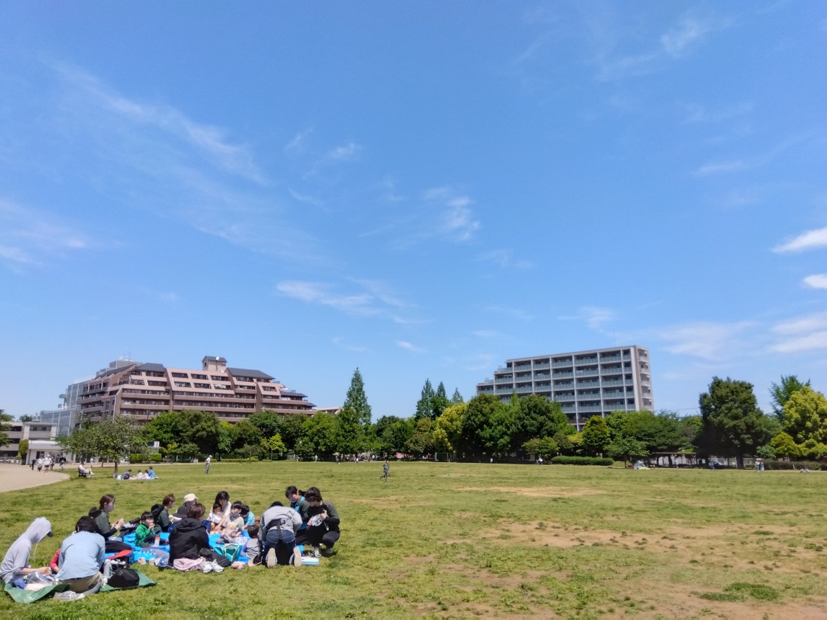 青空の下、草原にレジャーシートを広げ、大人と子どもたちがご飯を食べたり話をしたりしている。