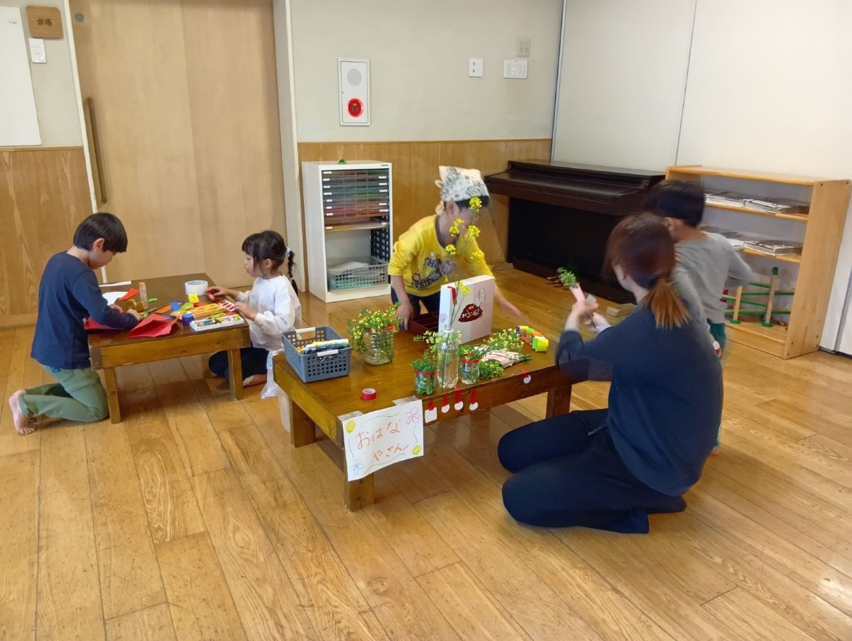 摘んできた草花はペットボトルの花瓶に活け、保育室内の「おはなやさん」と書かれた机の上に置かれている。机の傍で大人が花束を作り、子どもたちは話をしている。奥の机では、画用紙を折る子、クレヨンを取り出す子がいる。