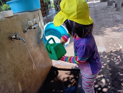 子どもが水道場に行き、じょうろに水を汲もうとしている。