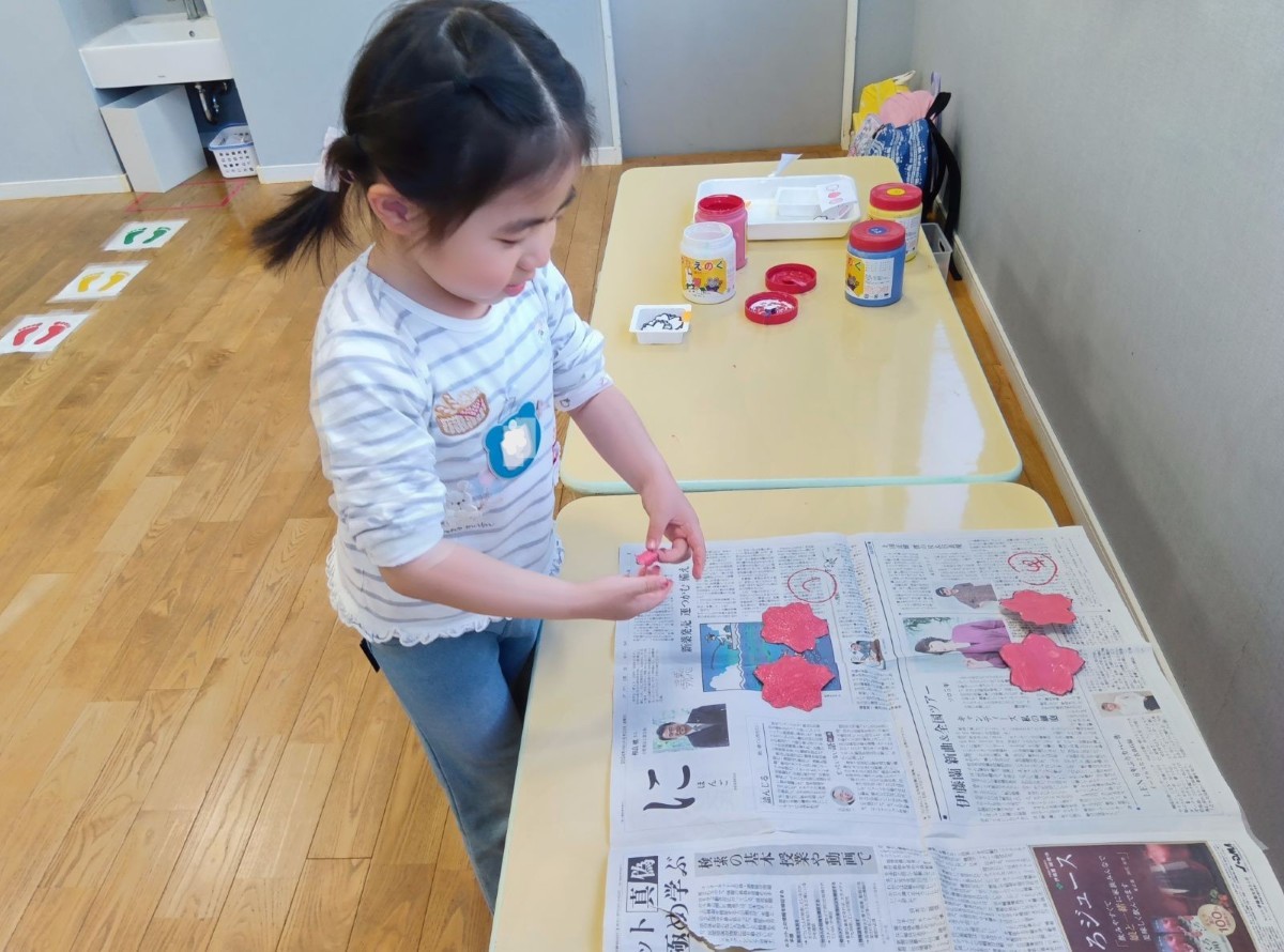 絵具、パレット、花の形にかたどられた画用紙が置かれている机と、色が塗られて新聞紙の上で乾かしている画用紙の花があり、子どもは色付けをした画用紙の花を手に持っている。