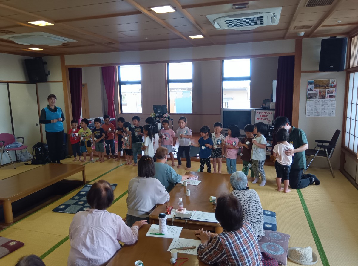 子どもたちは畳の和室で高齢者が座って見守る中、前に一列で並び、手遊びを行っている。子どもたちの両端に大人もいる。