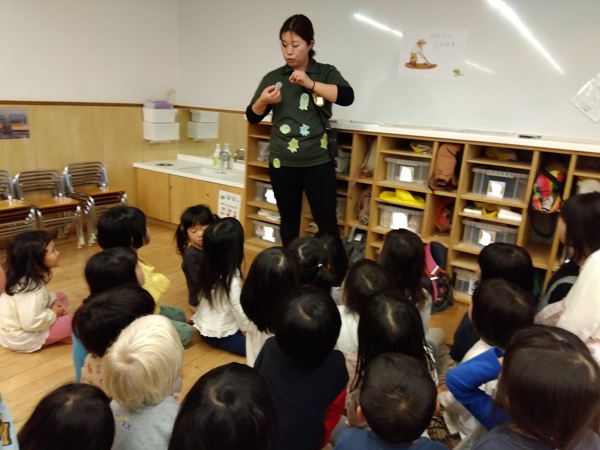 一人の大人が子どもたちの前に立ち、小さい画用紙を体に貼って見せ、子どもたちは座ってその様子を見ている。