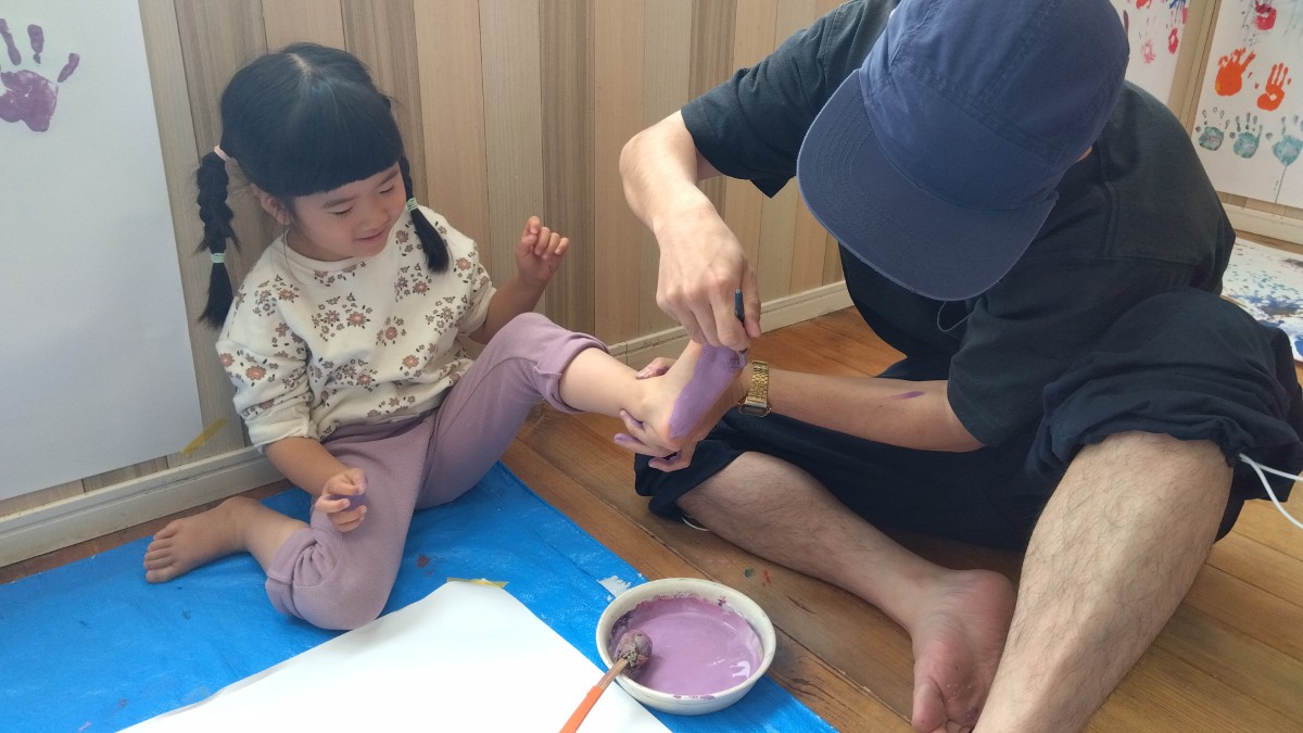 大人が筆を持って子どもの足裏に絵の具を塗っている。足元にはレジャーシートの上に画用紙が置かれている。