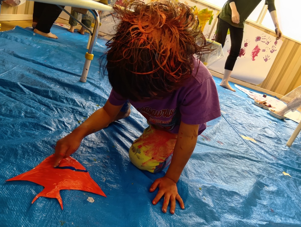 髪の毛やズボンに絵の具を付けた子どもが、絵の具の塗られた魚の形の画用紙に指を使って青い線を描いている。