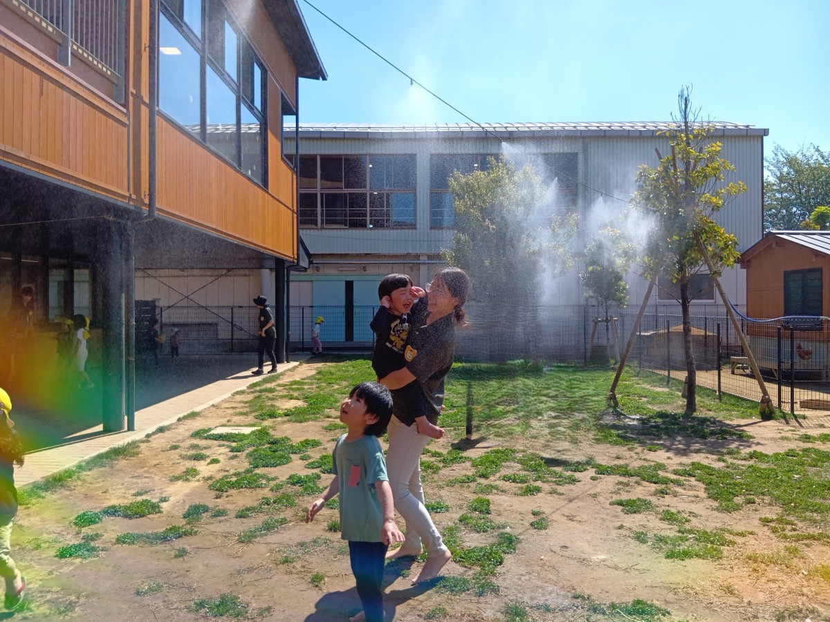 園庭ではミストの水しぶきが園庭に舞い上がり、上空を見上げる子どもや抱っこされている子どもが笑顔で顔をぬぐったりしている。