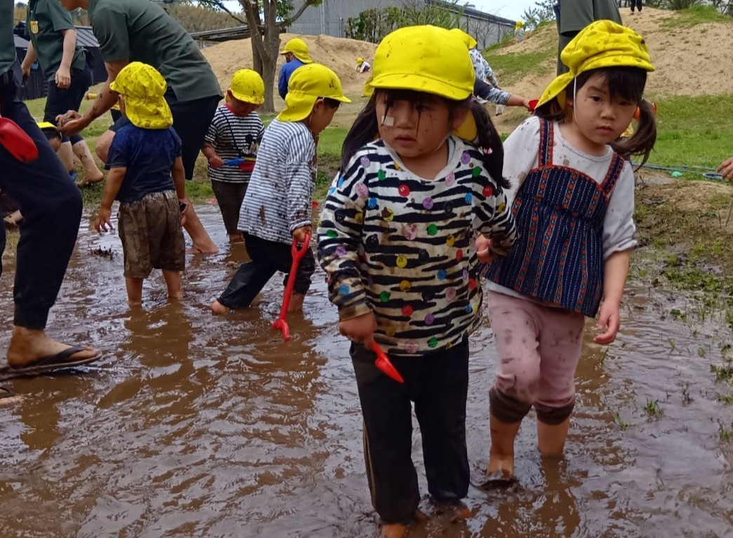 水が貼られた田んぼの中で、シャベルを持った子どもたちが泥だらけになって歩いている。