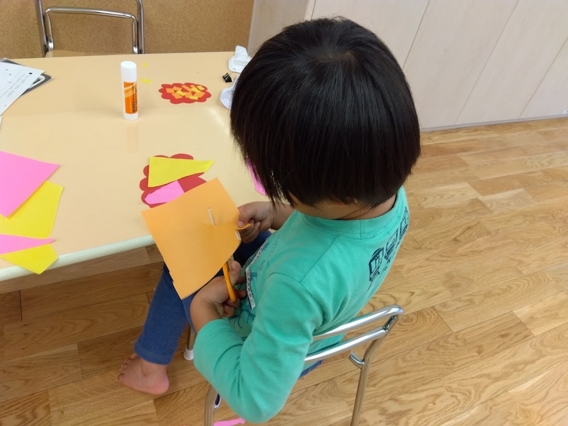 一人の子どもが椅子に座ってはさみで画用紙を切っている。