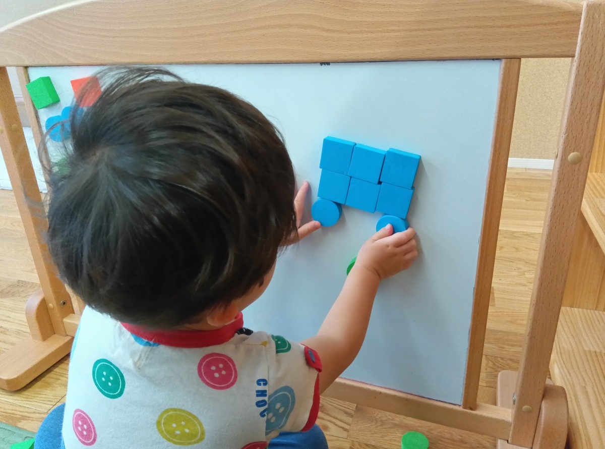 一人の子どもがマグネットボードに向かって四角と丸の形のブロックを張り付けて自動車の形を作っている。