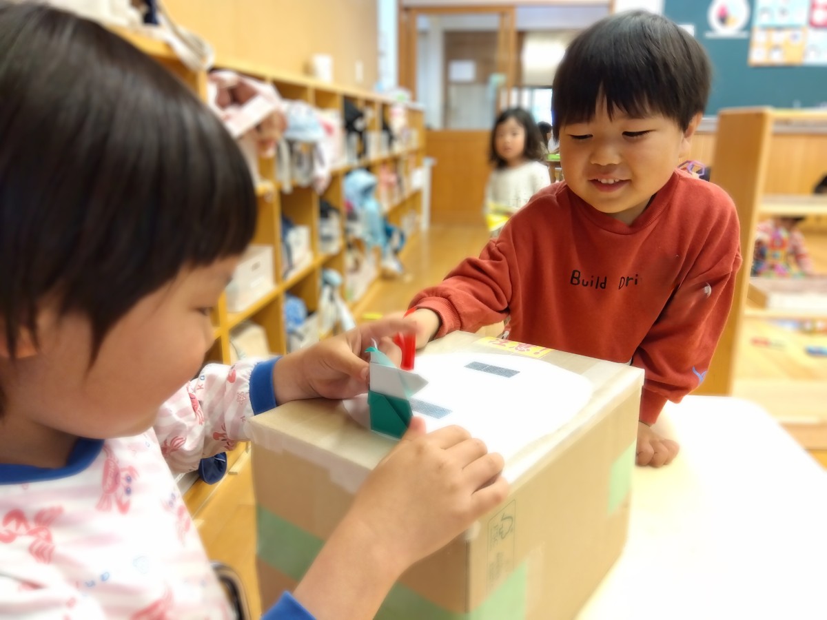 二人の子どもが、机の段ボールで作られた土俵に、折った折り紙を置いて段ボールを叩いている。