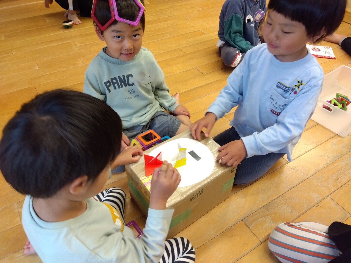 床に段ボールで作った土俵を置き、二人の子どもが向かい合って折り紙の乗った土俵を指先で叩いている。