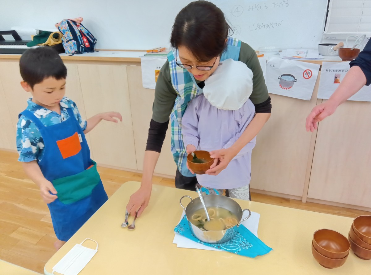 室内では、エプロンを着けた大人が一人の子どもの後ろに立ち、子どもが両手で持っている汁の入ったお椀を片手で支え、反対の手でスプーンを取ろうとしている。