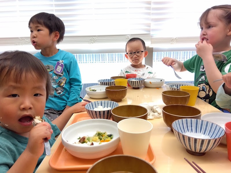 子どもたちは、同じ机でスプーンやフォークを使って食事を食べている。