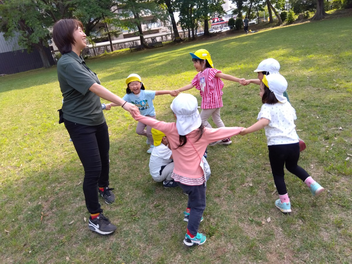 一人の子どもがしゃがみ、その周りを大人と多くの友達が輪になって手をつないで歩いている。