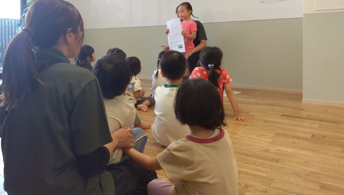 一人の子どもが一人の大人の膝の間に立ち、一枚の紙を持っている。多くの子どもたちはその様子を座って聞いている。