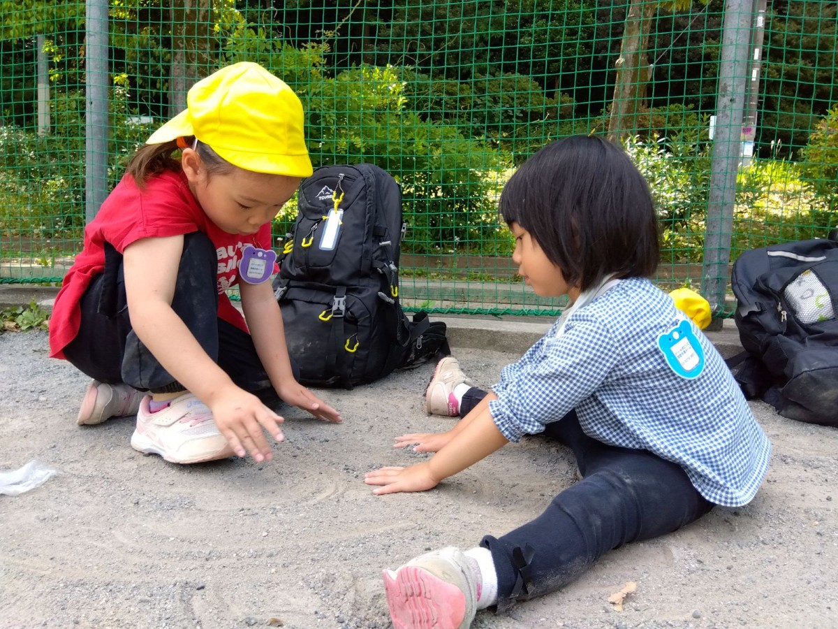 二人の子どもが向かい合って地面に座り、砂を触っている。後ろには、散歩リュックが置かれている。
