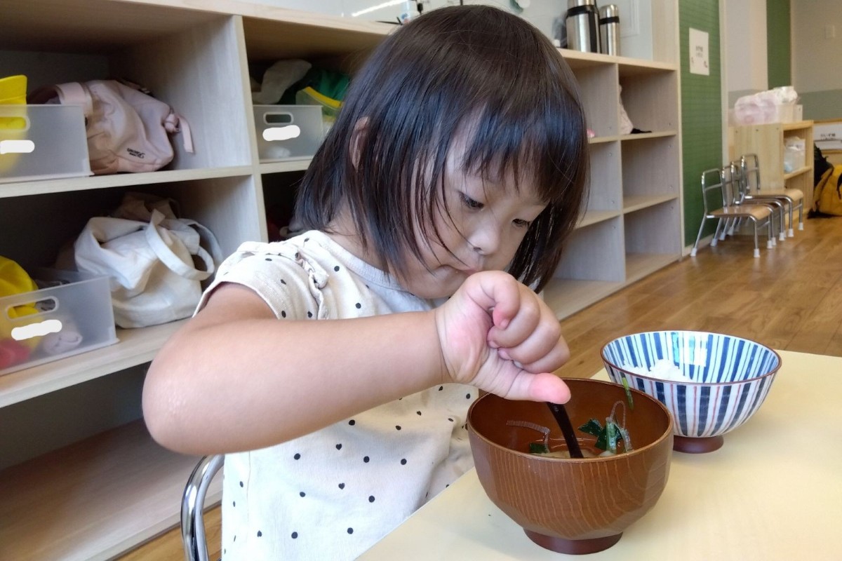 室内では、一人の子どもが上手持ちで食具を持ち、給食の汁物の具を掬おうとしている。