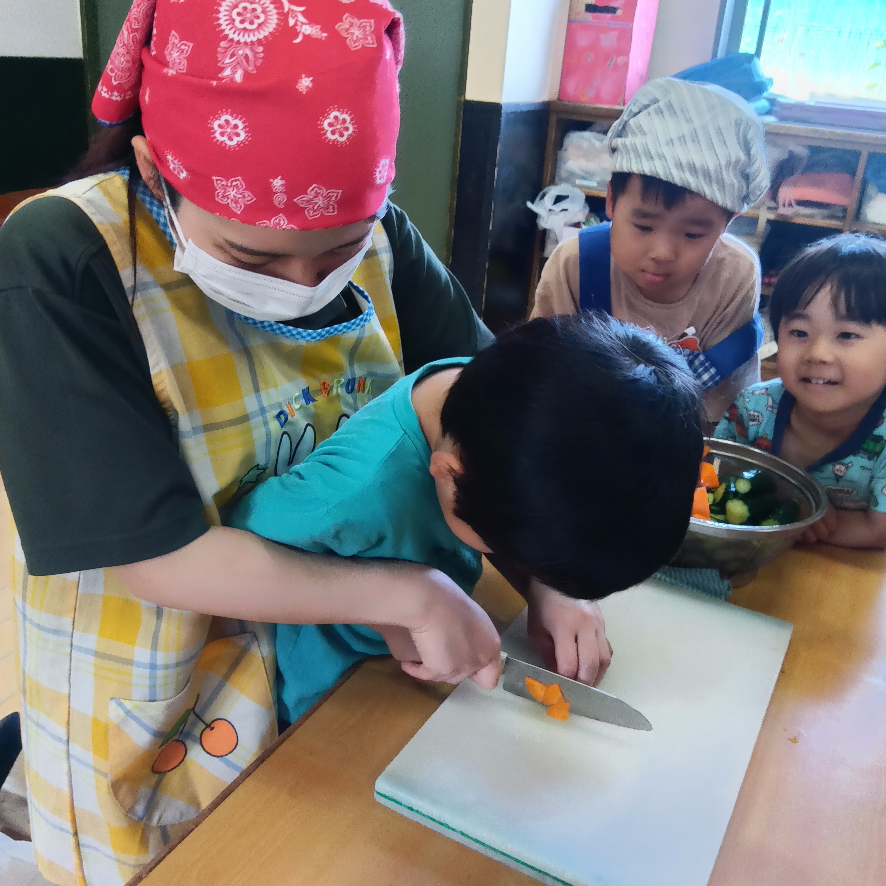 大人に後ろから手を添えられた一人の子どもが、ニンジンを包丁で切っている