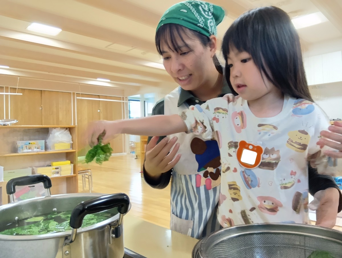 一人の子どもが、湯気の上がる鍋に小松菜の葉を入れようとしている。大人は子どもの後ろで見守っている。