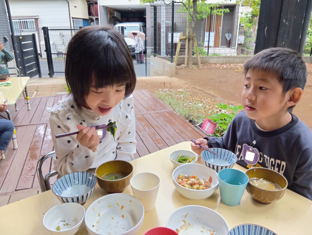 テラスでは、子どもたちが給食を食べており、一人の子どもが大きな口を開けて味噌汁に入った小松菜を食べようとしている。