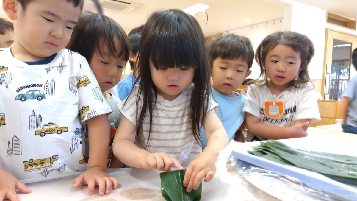 机の上にはたくさんの笹の葉が置いてあり、笹を巻いている一人の子どもの周りには、多くの子どもたちが集まってその様子を見ている。