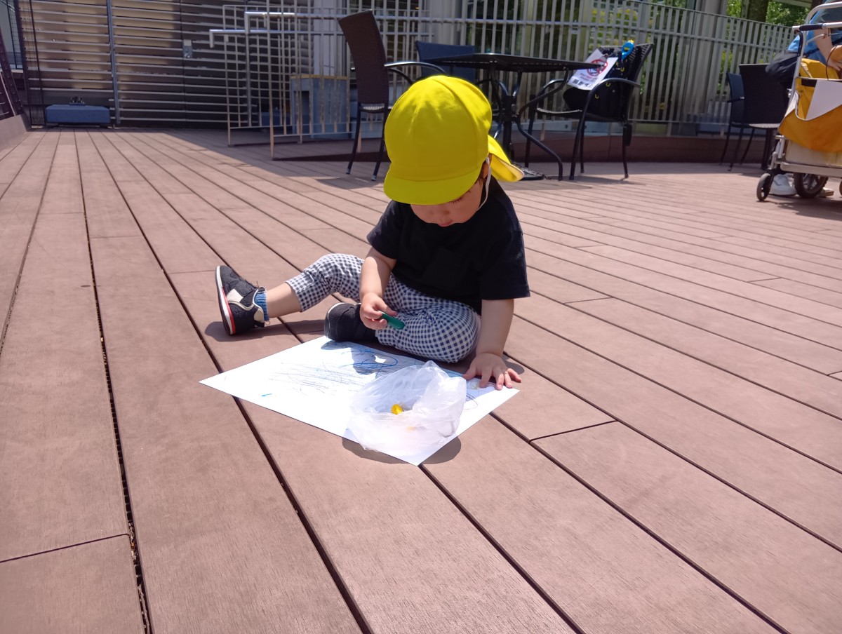 青空の下、一人の乳児が床に座り、画用紙になぐり書きをしている。