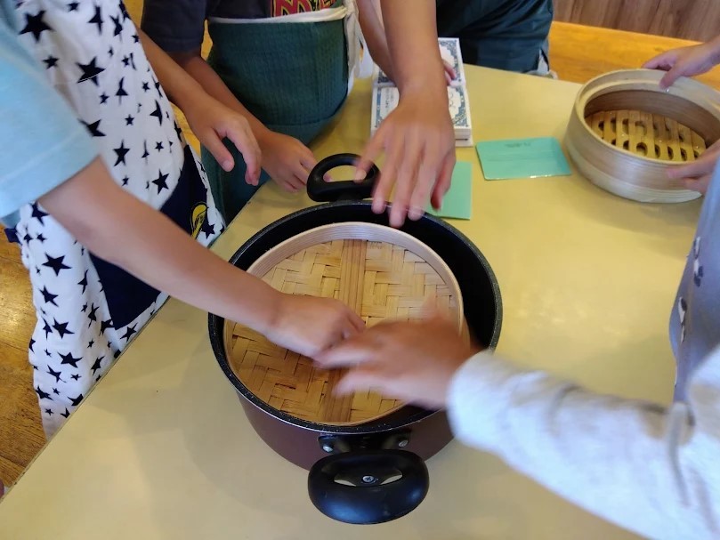子どもたちが集まる机の上には、蒸篭の蓋が入った鍋と、鍋の外に蒸篭の本体が置かれている