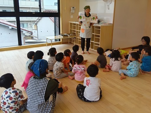 エプロンと三角巾を着けた一人の大人が前に立ち、座っている子どもたちに２枚のイラストを見せている。