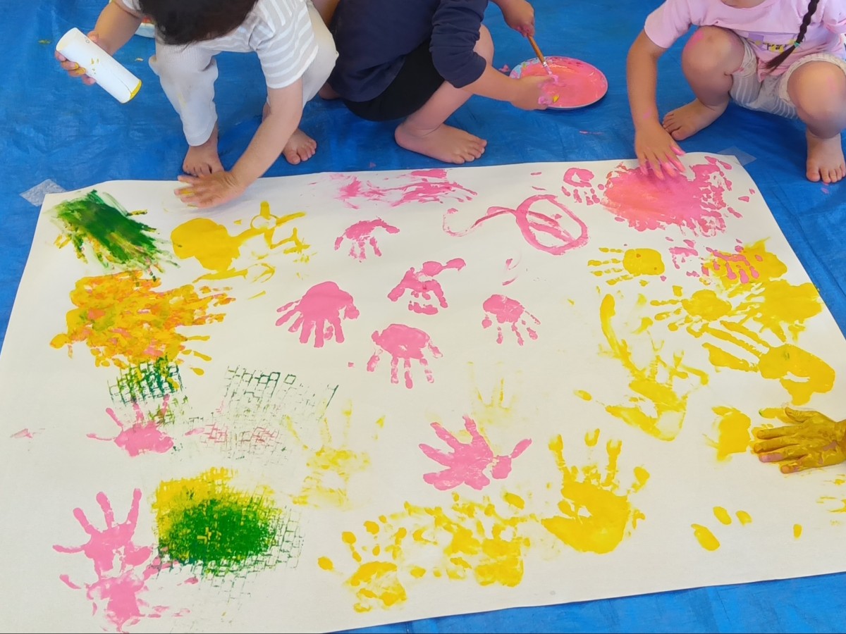 友達に掌に塗ってもらった絵の具で大きな模造紙に手形をつけている子、自ら手形を着ける子、ペーパー芯を片手に持って手形をつける子などがおり、模造紙は黄色や桃色で手形やローラーなどの模様ができあがっている。