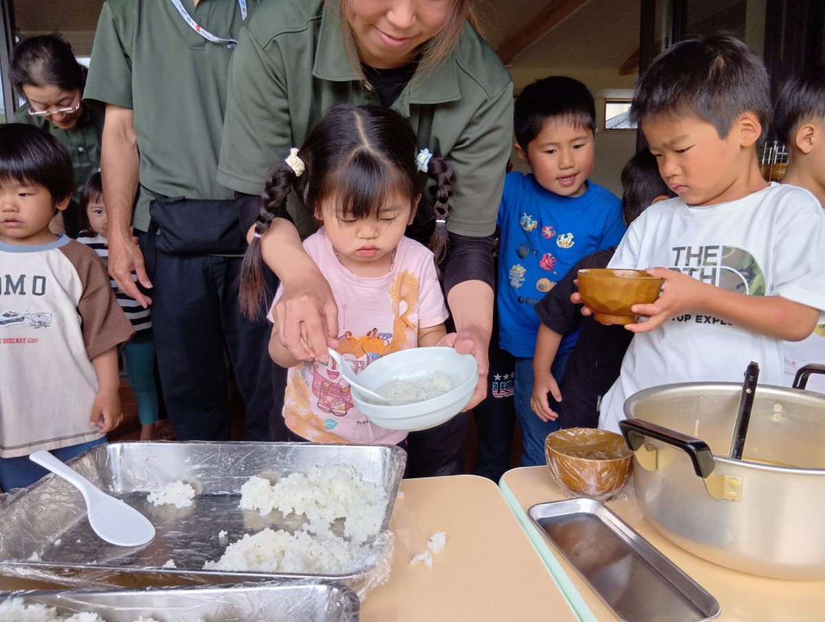 大人が子どもの後ろから手を添えて、ご飯を皿によそうことを手伝っている。