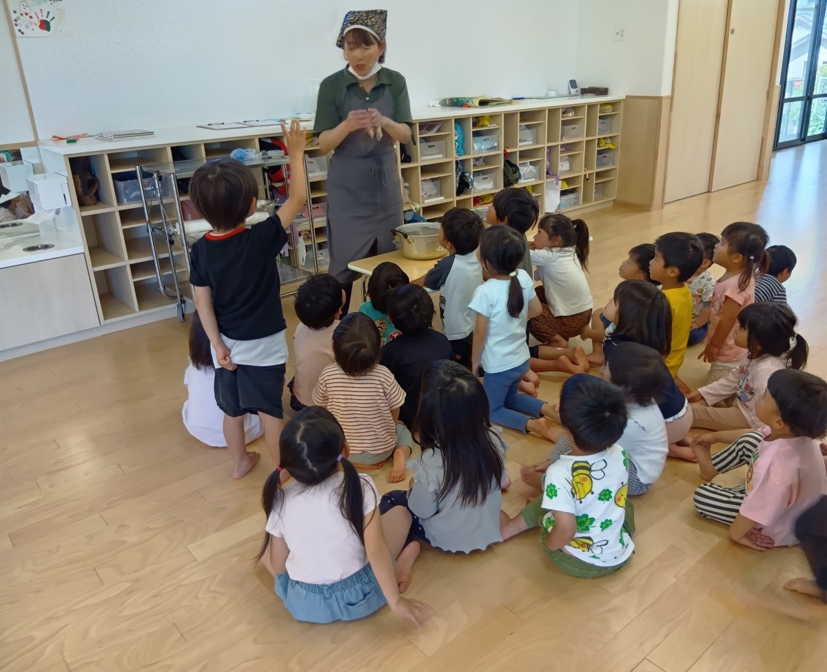 一人の大人が大きな鍋が置かれた机の前に立ち、座っている子どもたちに話しかけている。一人の子どもが立ち上がって手をあげている。