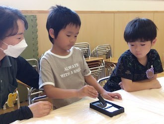 一人の子どもが机で背筋を伸ばして墨をすっており、その隣で大人と友達がその様子を見ている