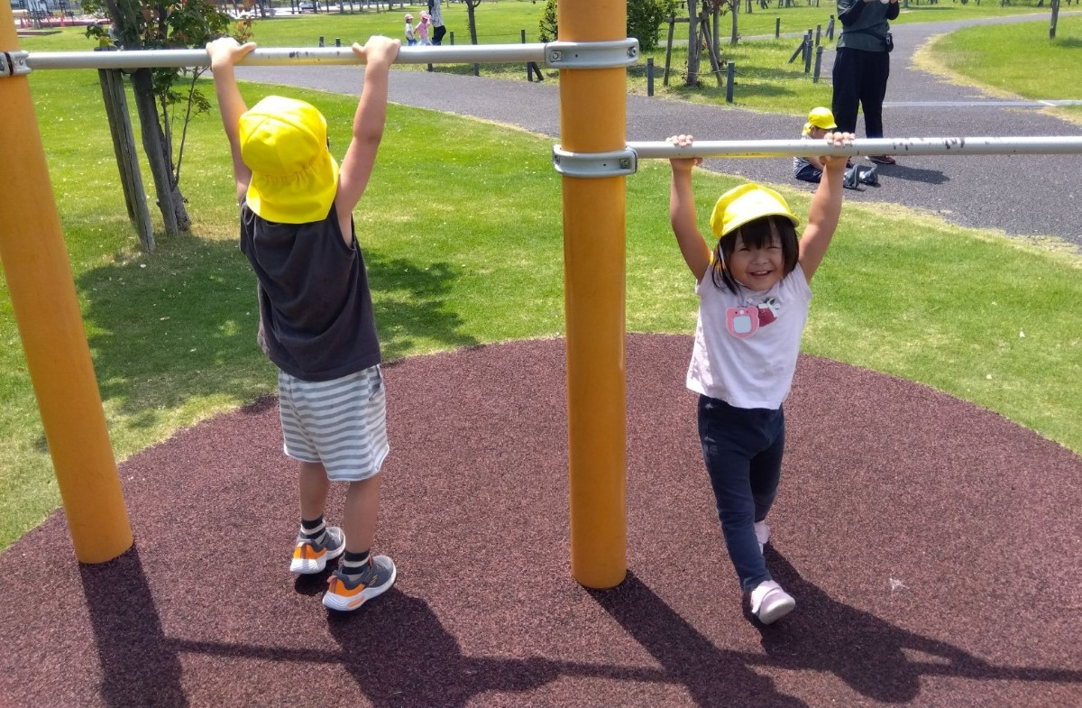 公園の鉄棒では、二人の子どもがぶら下がっており、一人はカメラに向かって笑顔を見せている
