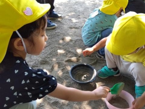 公園で、シャベルを持った子どもが、正面にいる友達の砂の入ったバケツの中に砂を入れている