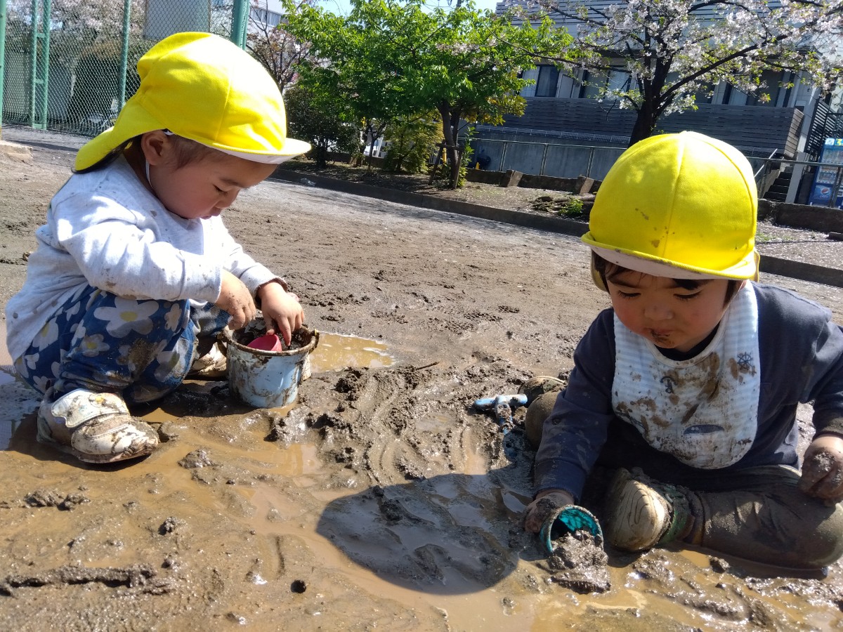 二人の子どもが泥の中に座り、カップやバケツで泥をすくっている