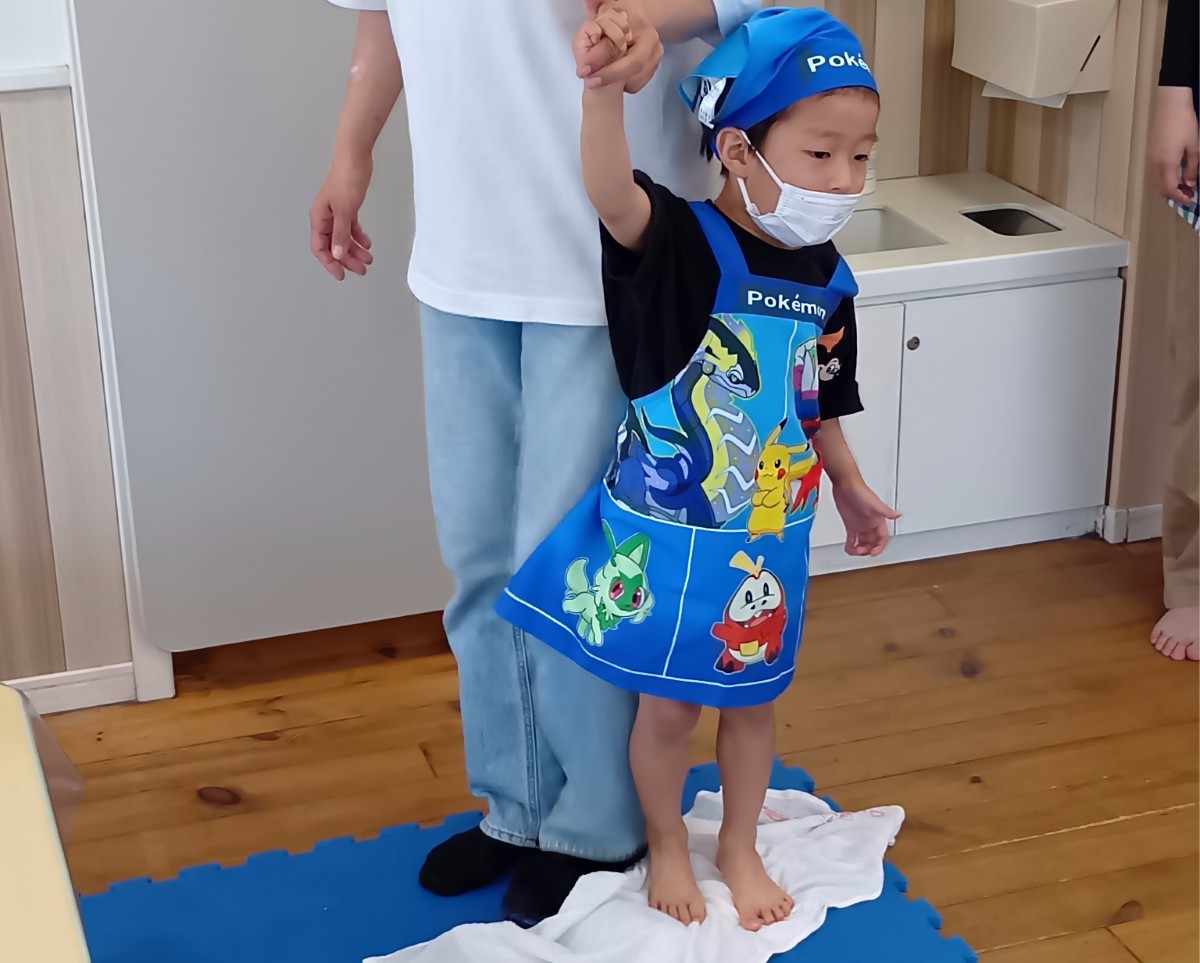 大人と手をつないだ子どもが床の上に置かれタオルが掛けられたうどんの生地を足で踏んでいる