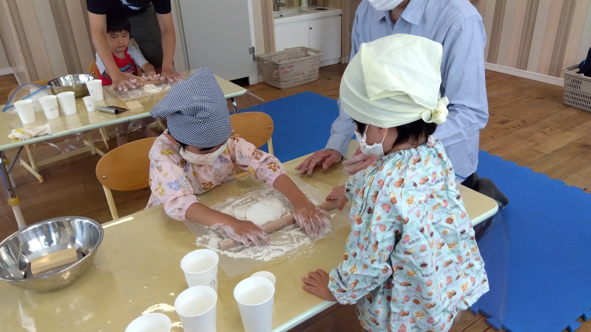 机が2台あり、それぞれの机で大人と一緒に子どもがうどんの生地を伸ばしたり、一人の子どもが生地を伸ばしその様子を大人ともう一人の子どもが近くで見ていたりする