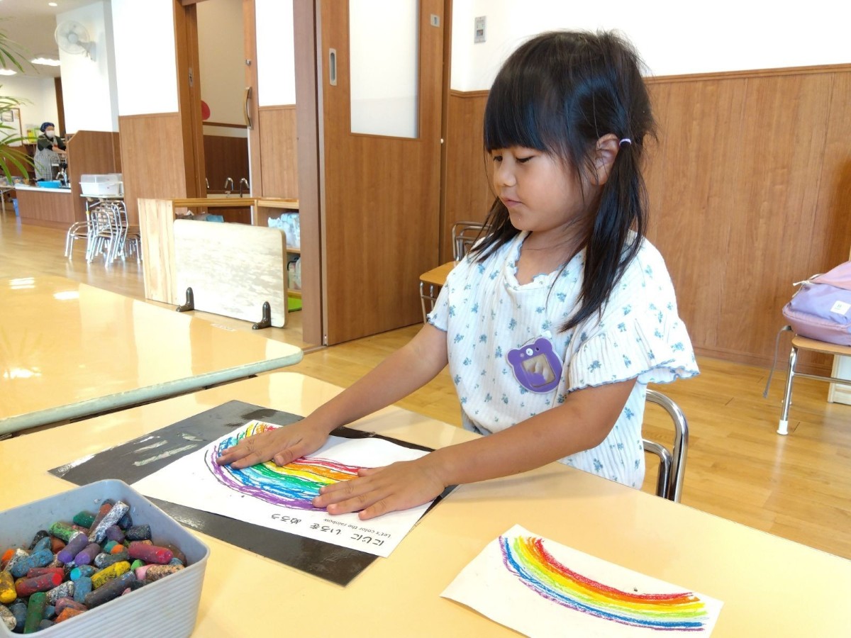 一人の子どもが虹を色鮮やかにクレヨンで描き、描いたクレヨンの上を掌で触っている