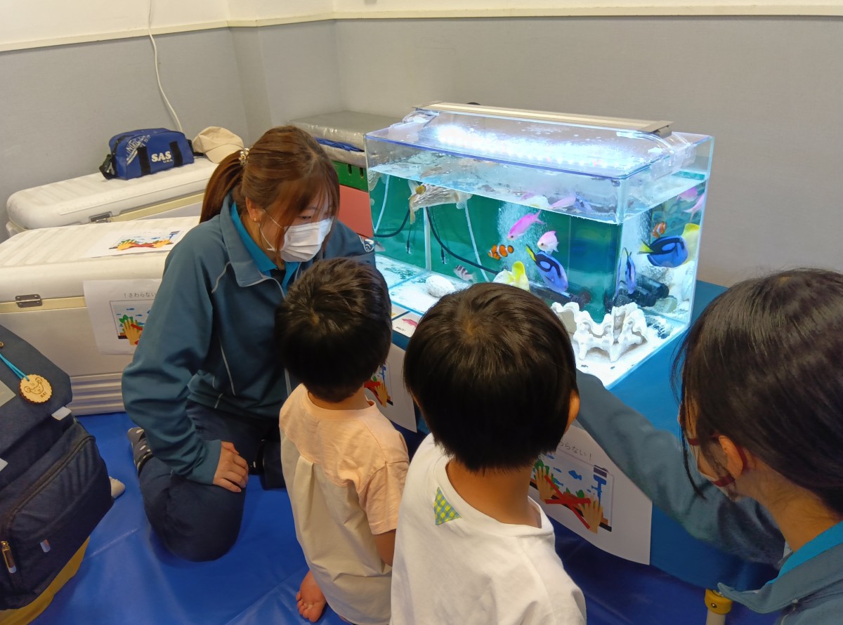熱帯魚が入っている水槽を座って近くでみる子どもたちと子どもたちに話しかける大人