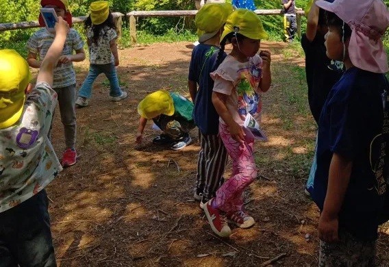 スタンプラリー台紙を首にかけた子どもたちが公園の林の中で地面や上空をみて探し回っている