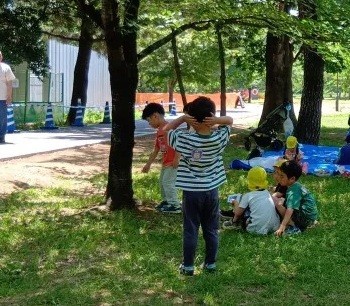 公園では、シートの上で座っている子、数名で集まって座っている子、立ってその様子を見ている子などがいる