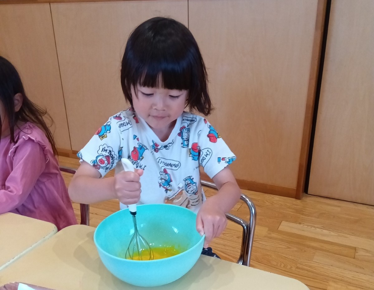 一人の子どもが片手に泡だて器を持ち、反対の手でボールを抑えてボールの中の卵を混ぜている