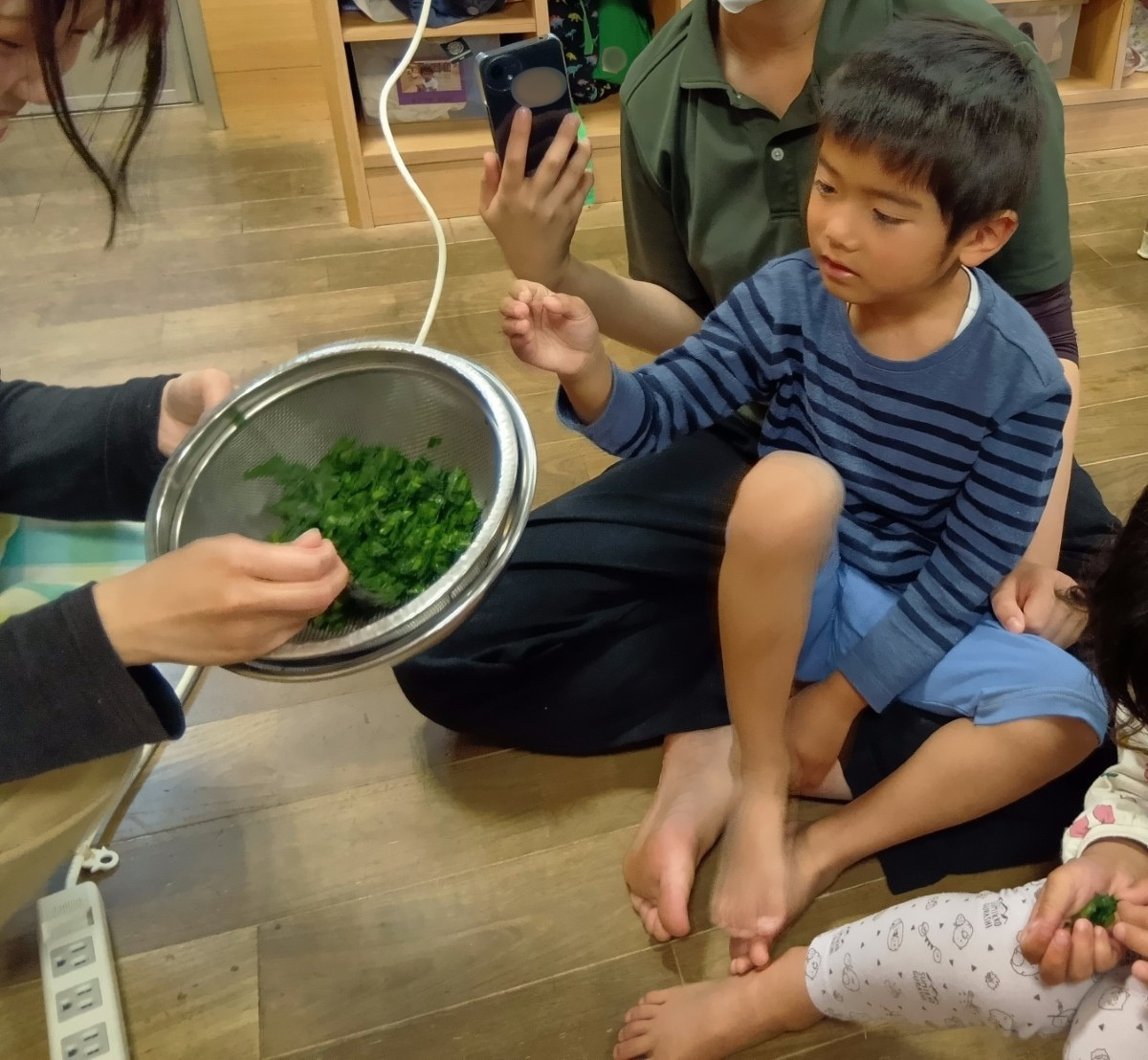 ざるの中に小口切になった葉物野菜が入っており、大人がその野菜を座っている子どもたちに渡そうとしている