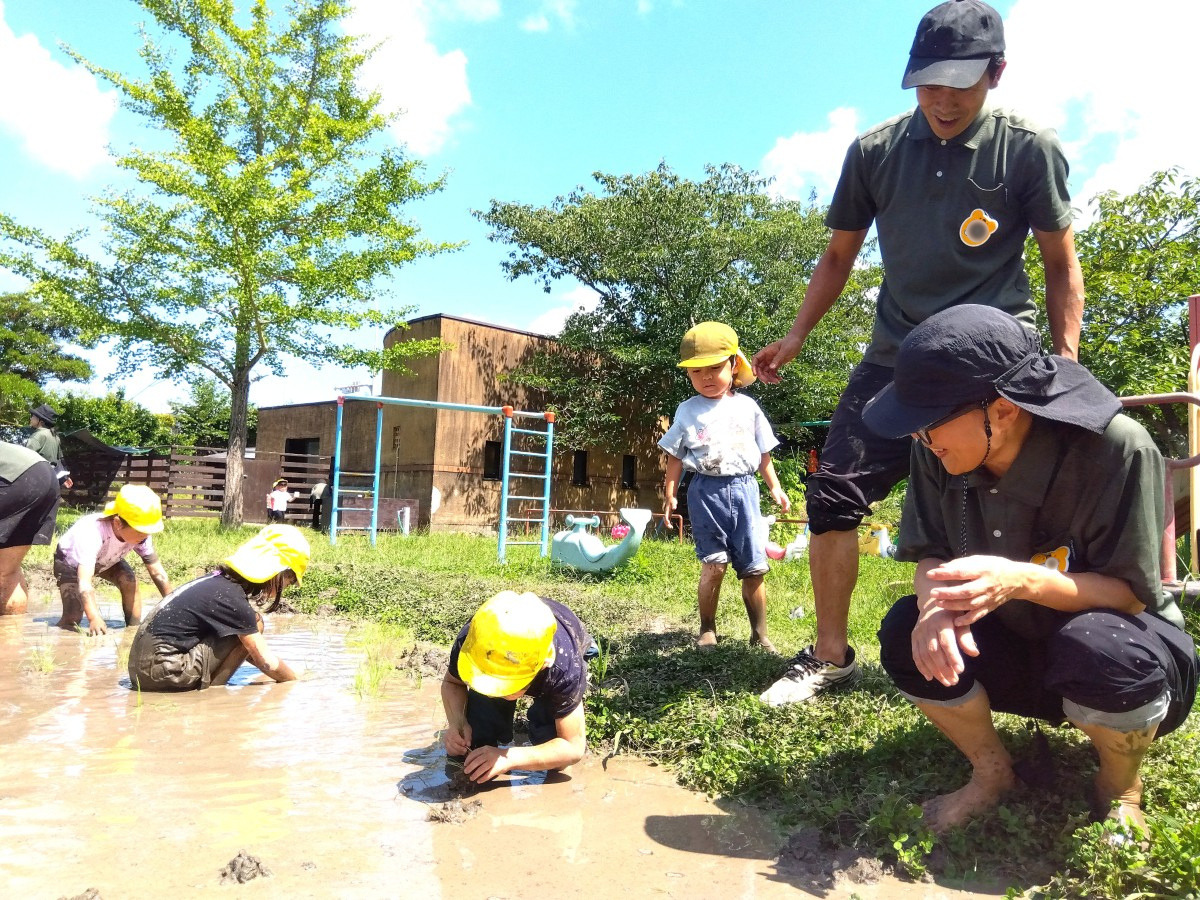 一人の子どもがかがんで田んぼの中をのぞいており、その様子を大人たちが笑顔で見守っている